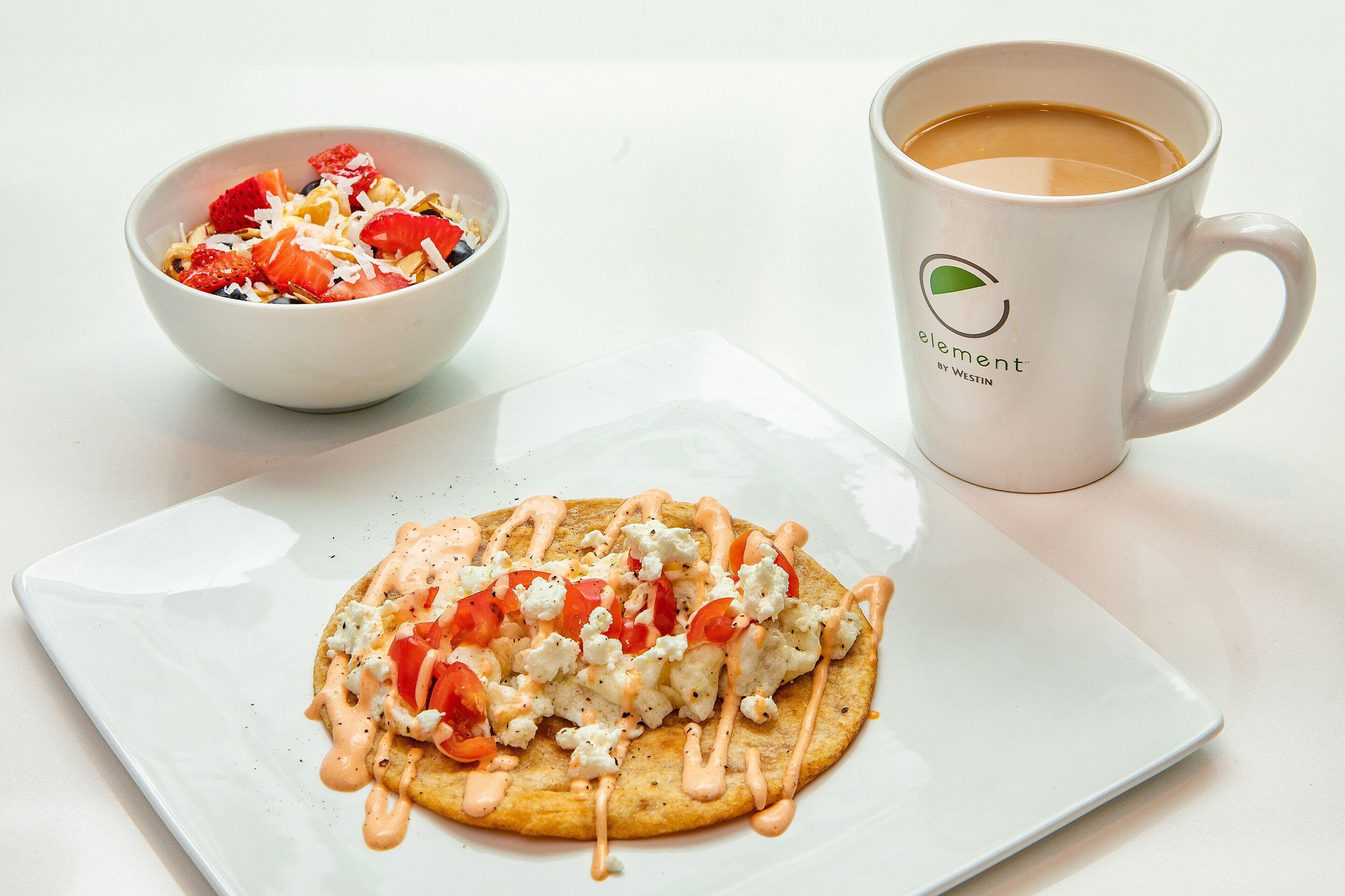 Un plato blanco con un alimento cubierto de tomates, queso y salsa, un bol blanco con ensalada de frutas y una taza blanca con un logotipo.