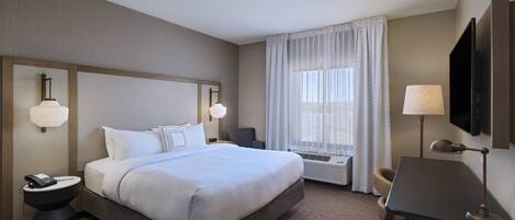A hotel room with a large bed, bedside tables, a desk, a chair, a TV, and a window with curtains.