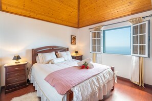 Un dormitorio con techo de madera, una cama grande con ropa de cama blanca, una mesita de noche con lámpara y una ventana con cortinas.