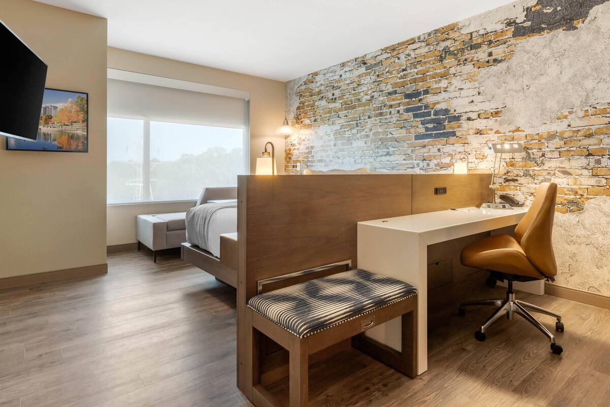 Ein modernes Zimmer mit Schreibtisch aus Holz, einem gepolsterten Sessel, einem Bett und einer Ziegelwand.
