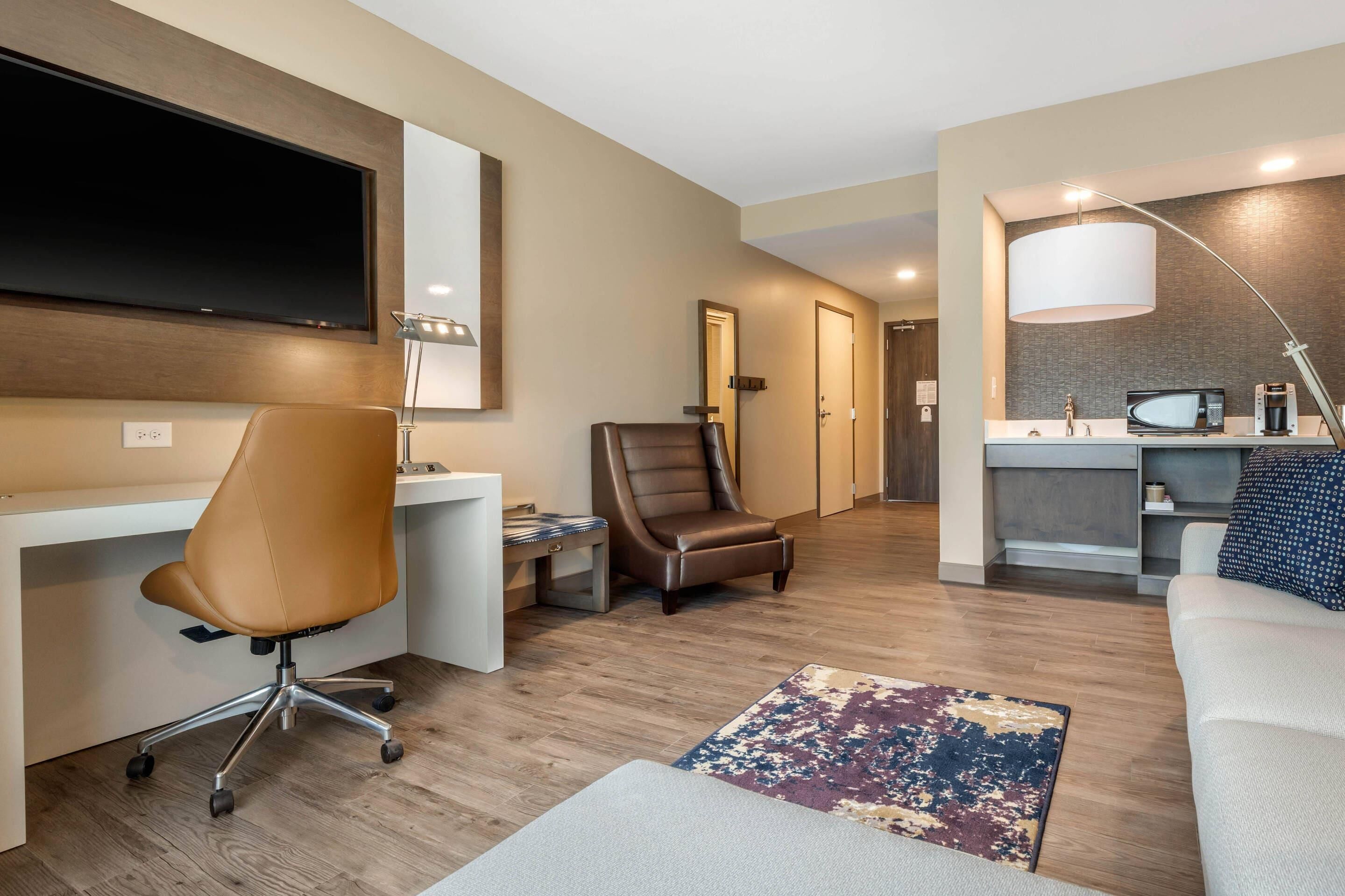 A modern hotel room with a desk, office chair, leather armchair, and a living area with a sofa and a patterned rug.