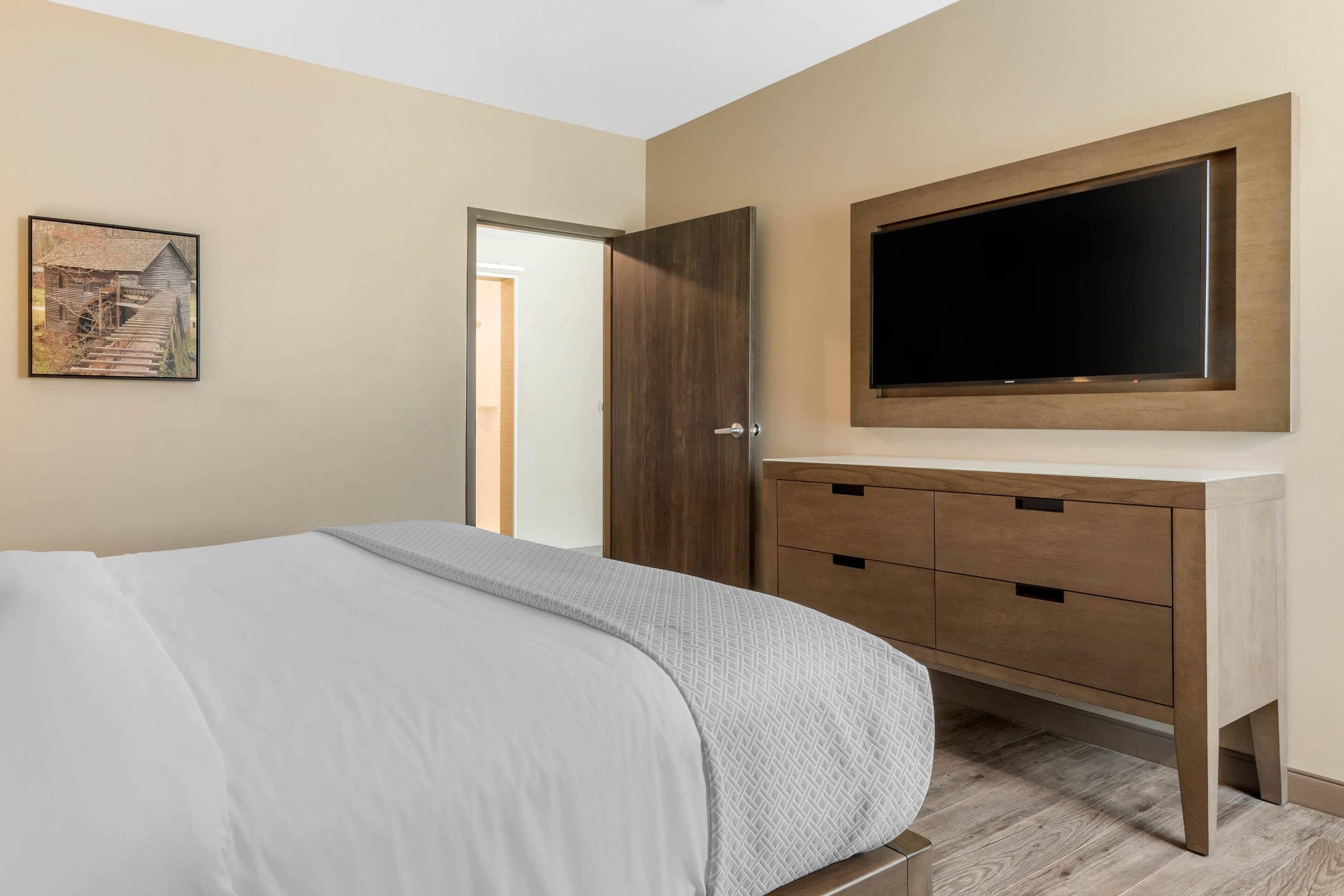 Une chambre à coucher avec un lit, une commode en bois, un téléviseur fixé au-dessus, et un tableau accroché au mur.