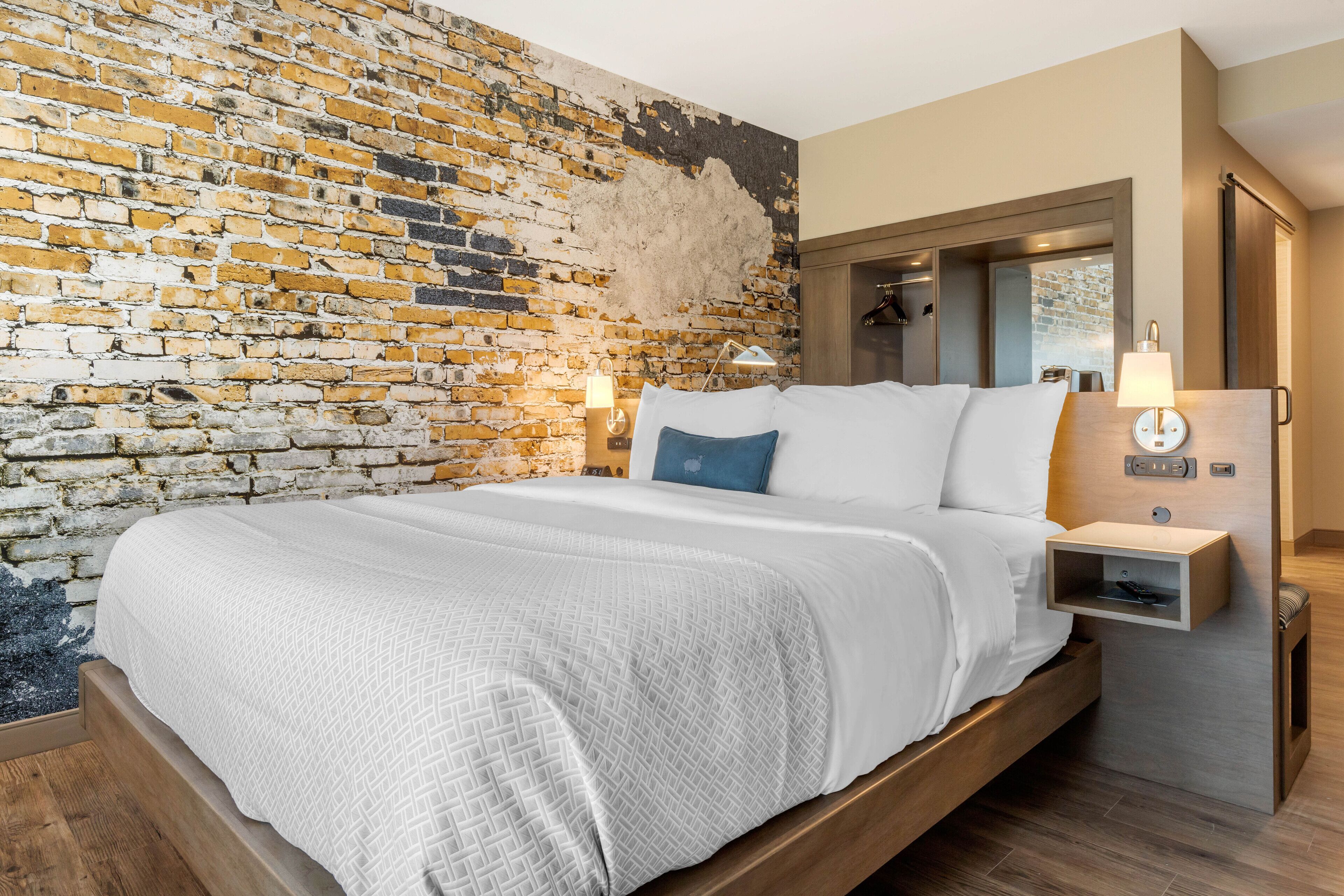 A modern hotel room with a brick wall, a wooden bed frame, and a nightstand with a lamp.