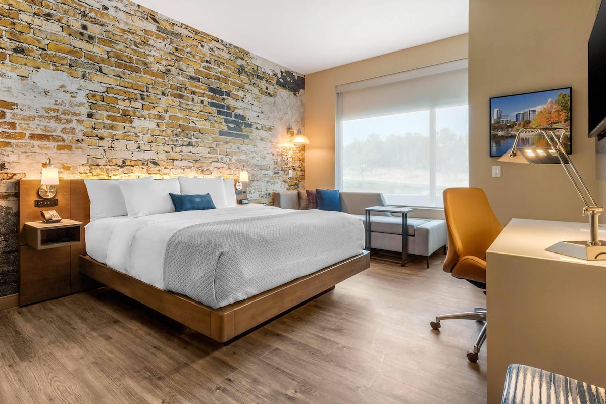 Ein modernes Hotelzimmer mit Backsteinwand, einem großen Bett, einem Schreibtisch und Blick ins Freie.