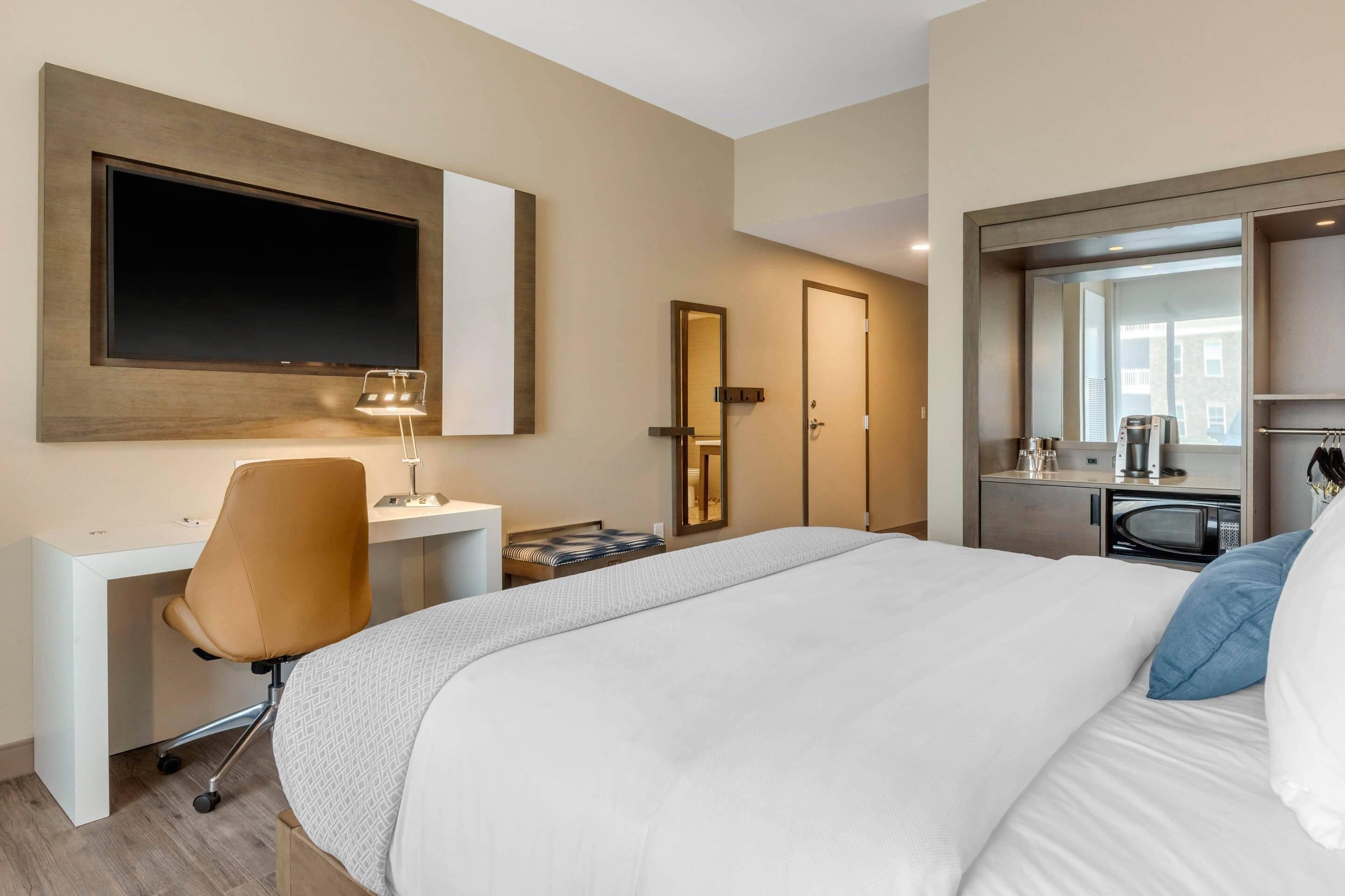 A hotel room with a large bed, a desk with a chair, a flat-screen TV mounted on the wall, a mirror, a wardrobe, and a kitchenette area.