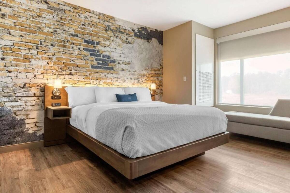 Ein modernes Schlafzimmer mit Backsteinwand, einem Bett mit weißer und grauer Bettdecke, einem Nachttisch mit Lampe und einem Fenster mit Blick.