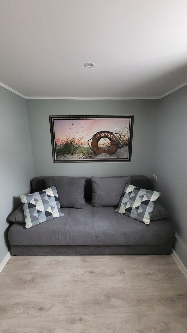 A grey sofa with two patterned cushions, a framed picture of a lifebuoy on a beach, and a recessed ceiling light.