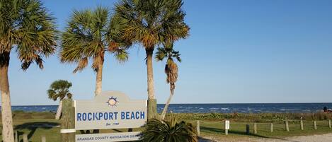 A sign for Rockport Beach with palm trees and the ocean in the background.