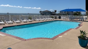 Una piscina con sillones blancos y un dosel azul, rodeada por una valla blanca y vistas al océano.