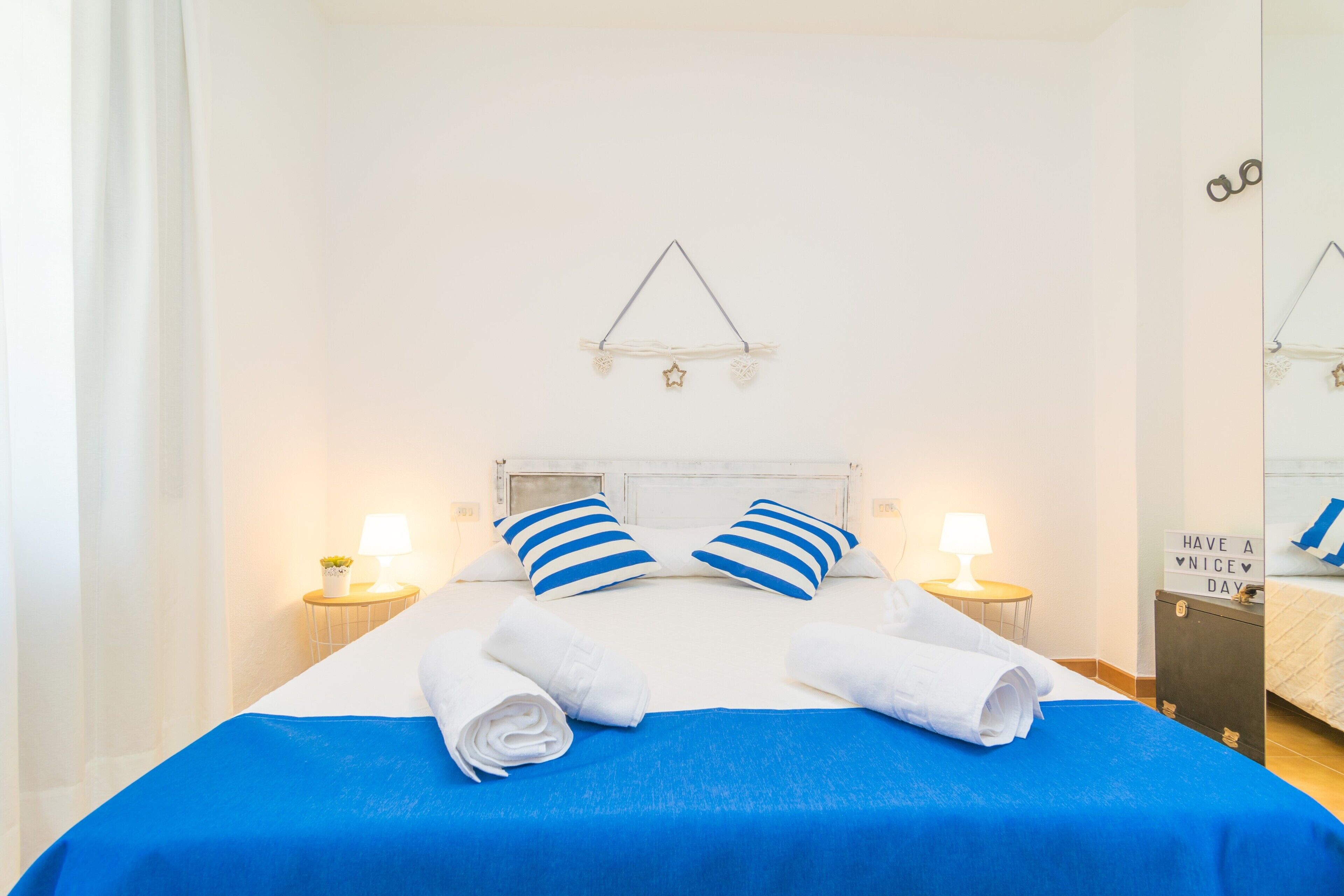 A neatly made bed with blue and white striped pillows, white towels, and a blue bedspread. There are two bedside tables with lamps, a wall-mounted light fixture, and a sign on the wall.