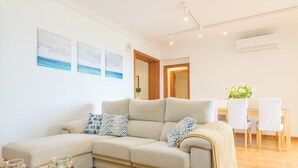 A modern living room with a beige sectional sofa, a glass coffee table, and a dining area with white chairs and a wooden table.