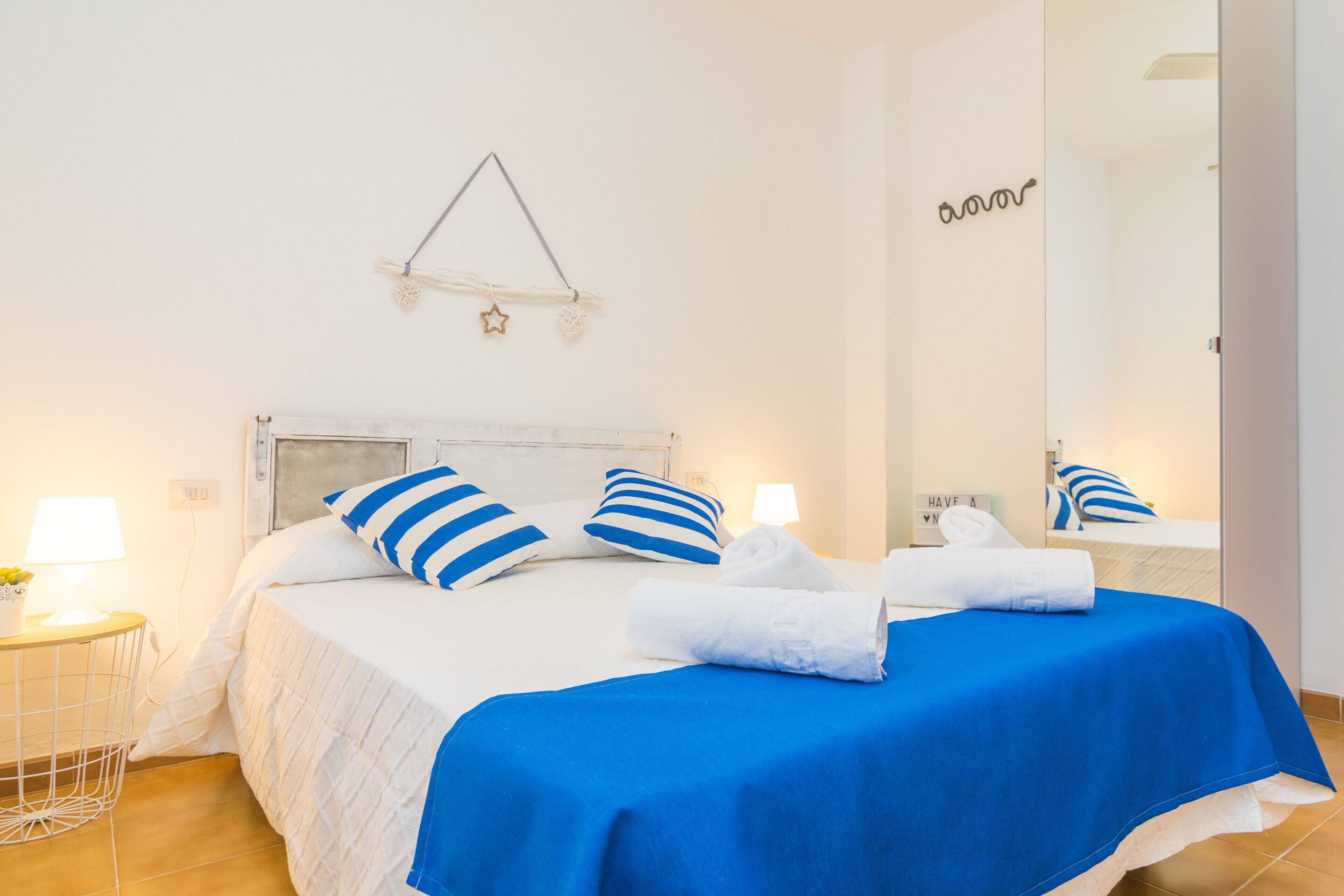 Una cama bien hecha con almohadas a rayas azules y blancas, un cabecero blanco y una colcha azul. Hay una mesita de noche con una lámpara y un objeto decorativo. Un espejo refleja parte de la habitación, y se observa una lámpara fijada a la pared.