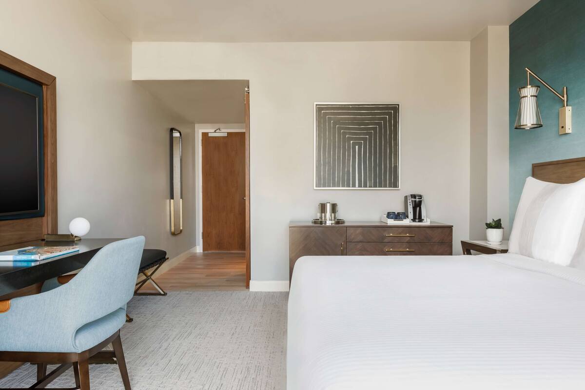Habitación de hotel con una cama grande, un escritorio con silla, una cafetera y una lámpara de pared.