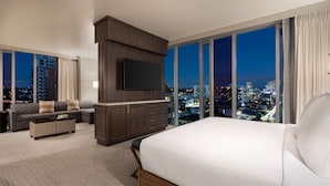 Habitación de hotel moderna con una cama grande, un televisor montado en un mueble de madera, un sofá y vistas a la ciudad por la noche.