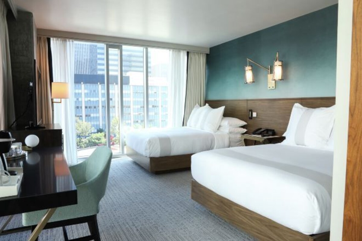 Habitación de hotel con dos camas, un escritorio, una silla, una lámpara y vistas a la ciudad.