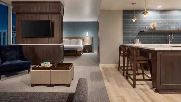 A modern living room with a large TV, dark wood furniture, a blue sofa, and a kitchen area with a bar and stools. - Carte Hotel San Diego Downtown, Curio Collection by Hilton (San Diego)