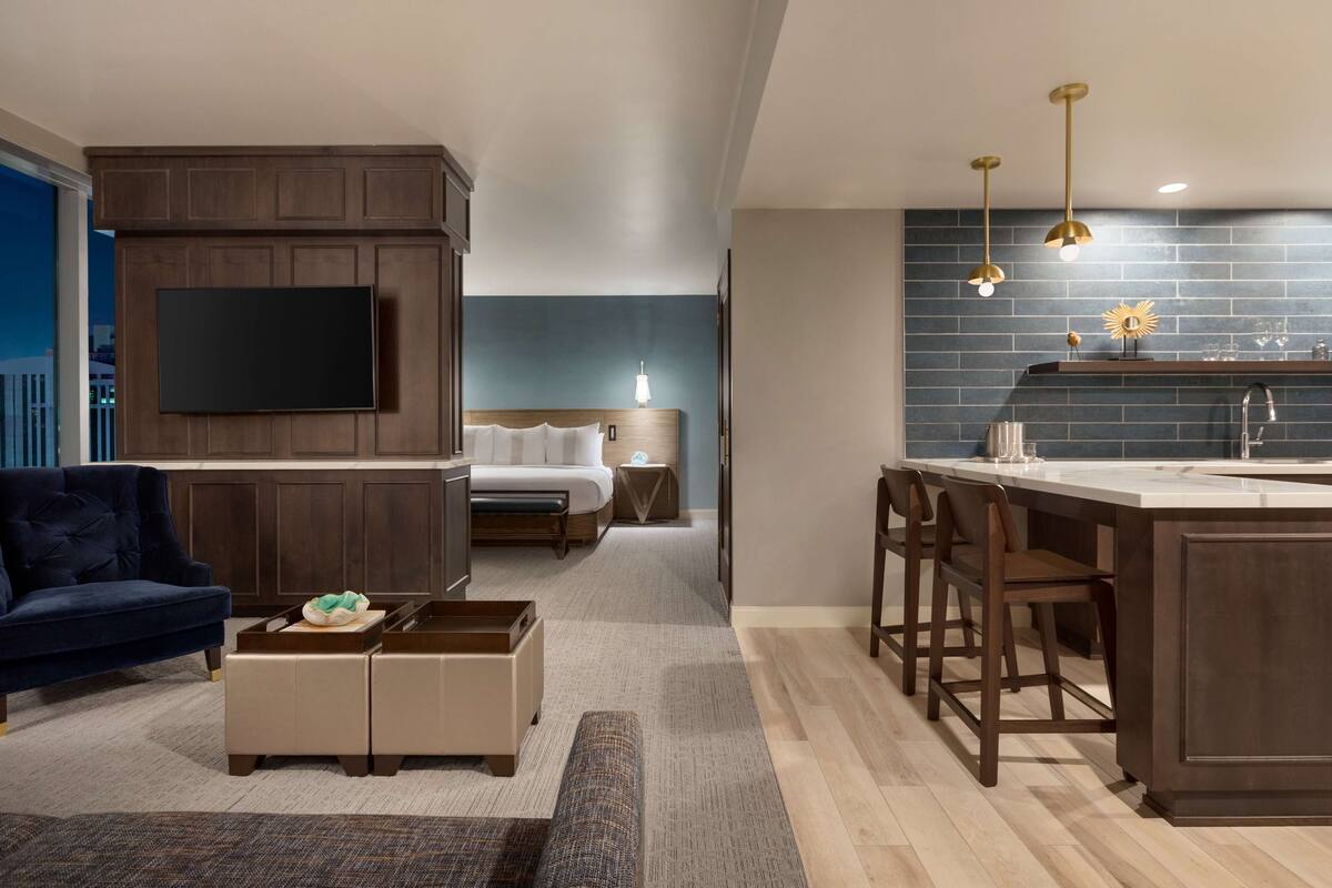 Una sala de estar moderna con un televisor grande, muebles de madera oscura, un sofá azul y una zona de cocina con barra y taburetes.