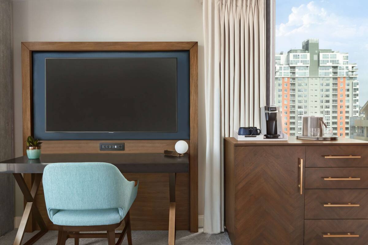 Habitación de hotel con televisor de pantalla plana grande montado en la pared, un escritorio de madera con una silla, minibar y vistas a edificios altos.