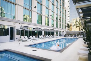 A swimming pool with lounge chairs and umbrellas, surrounded by a modern building with large windows. - Carte Hotel San Diego Downtown, Curio Collection by Hilton (San Diego)