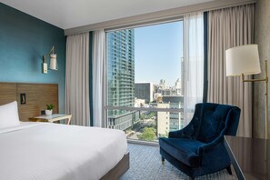 A hotel room with a large bed, a blue armchair, a small table with a plant, and a view of a cityscape through large windows. - Carte Hotel San Diego Downtown, Curio Collection by Hilton (San Diego)