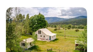 Une maison de campagne blanche avec une porte bleue, entourée d’une cour clôturée et d’un petit abri, se dressant sur un fond de champs verdoyants et de montagnes.