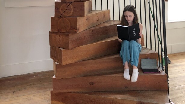 Un escalier en bois sur lequel est assise une jeune fille, qui lit un livre intitulé « Féministe ».