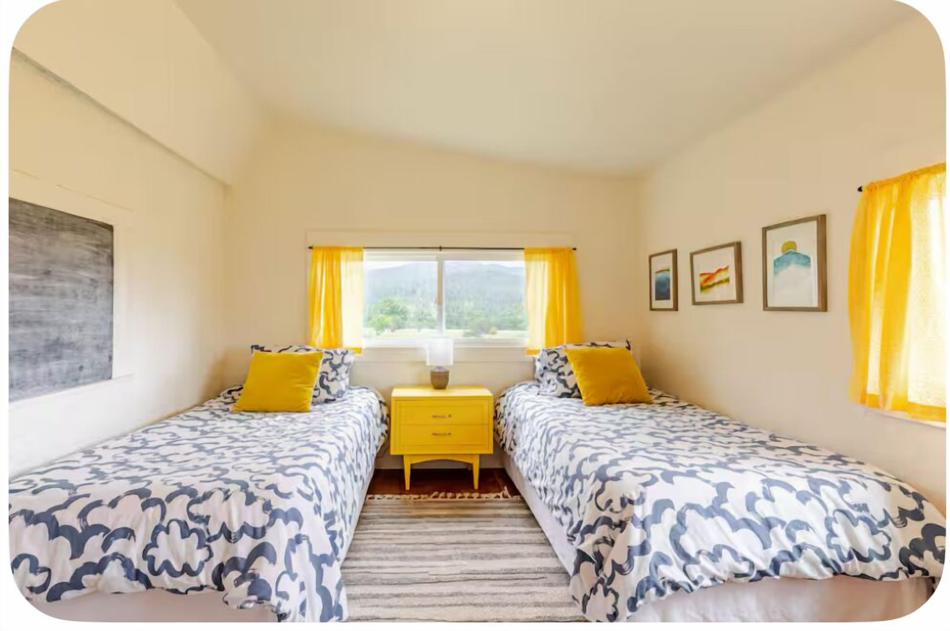 A bedroom with two beds, a nightstand, a window with curtains, and framed pictures on the wall.