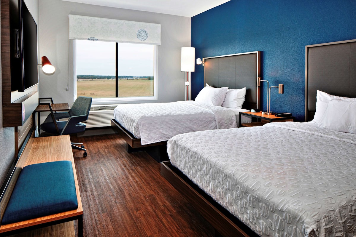 A hotel room with two beds, a desk, a bench, and a large window offering a view.