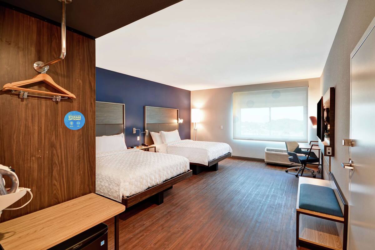 A hotel room with two beds, a desk, a chair, a TV, and a window with blinds.