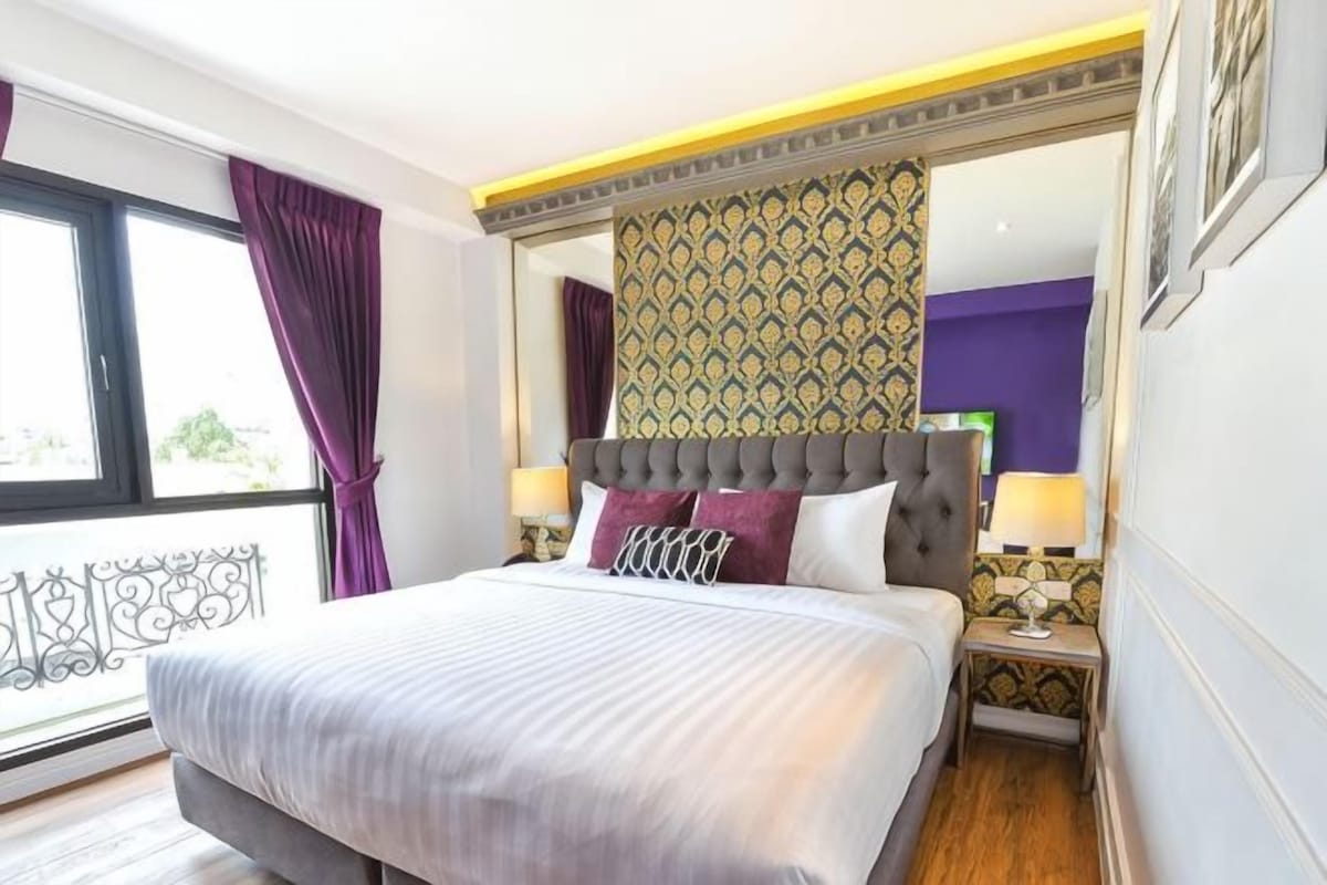 A hotel room with a large bed, a nightstand, a mirror, and a view of the outdoors.