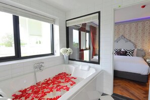 Une salle de bain moderne avec une baignoire, un grand miroir et une vue sur une chambre à coucher avec un lit et une table de chevet.