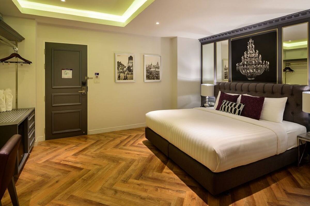 Une chambre avec un grand lit, un parquet en bois disposé en point de Hongrie, une porte avec un miroir et deux tableaux encadrés accrochés au mur.