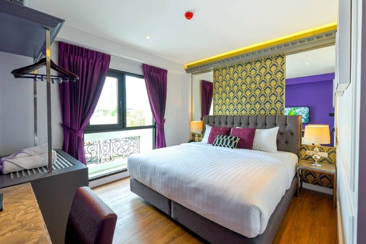 Une chambre d’hôtel avec un grand lit, un bureau avec une chaise, une fenêtre avec des rideaux violets et un mur à motifs.