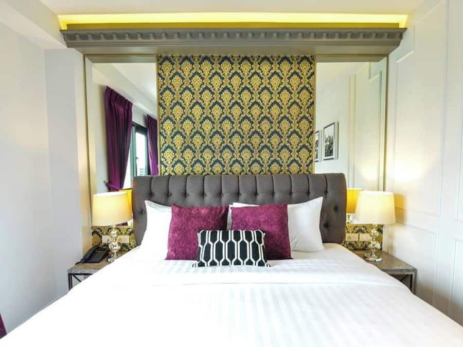Quarto de hotel com uma cama grande, duas mesas de cabeceira com abajures, cabeceira com papel de parede estampado e uma janela com cortinas roxas.