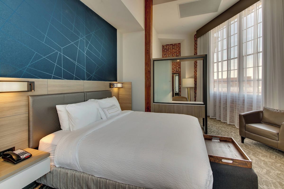A hotel room with a large bed, a nightstand with a phone, a bedside table with a lamp, a mirror, and a window with sheer curtains.
