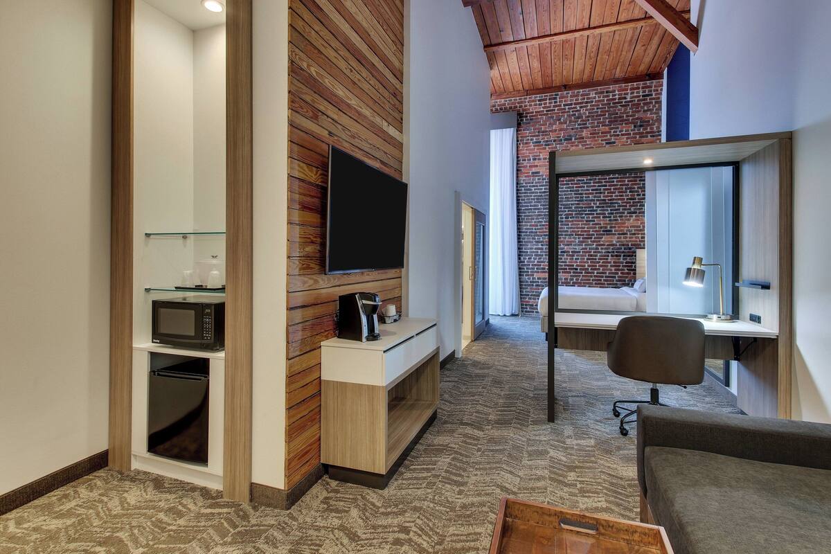 A modern hotel room with a wooden ceiling, a brick wall, a flat-screen TV, a microwave, a coffee maker, a desk, and a chair.