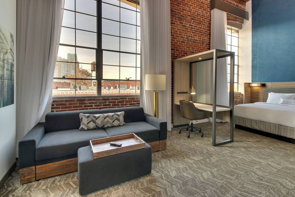 A modern hotel room with a large window, a sofa, a desk, and a bed.
