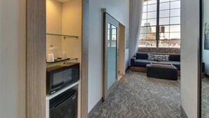 A narrow hallway with a microwave and a glass shelf, leading to a seating area with a sofa and a window with a city view. - SpringHill Suites by Marriott Montgomery Downtown (Montgomery)