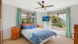 A bedroom with a bed, wooden nightstands, a ceiling fan, and large windows with curtains. - Kianga Parade Beach House (KIANGA)