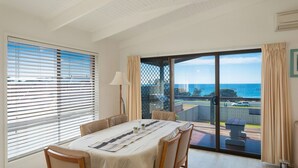 A spacious room with a large dining table, comfortable seating, and a view of the ocean through a sliding glass door. - Kianga Parade Beach House (KIANGA)