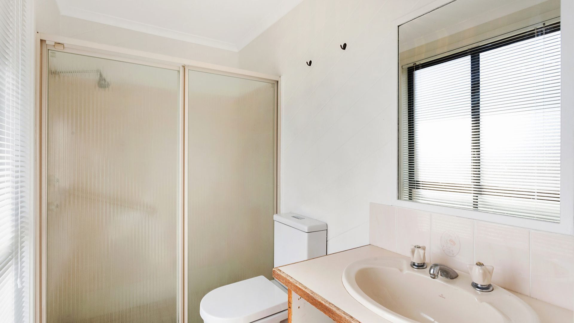 A bathroom with a glass shower enclosure, a toilet, a sink with a mirror, and a window with blinds.