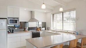 A modern kitchen with white cabinetry, a central island, a stove, a microwave, and a large window with blinds. - Kianga Parade Beach House (KIANGA)