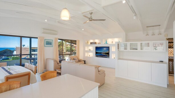 A modern living room with a large TV, a dining area, and a balcony with a view of the sea. - Kianga Parade Beach House (KIANGA)