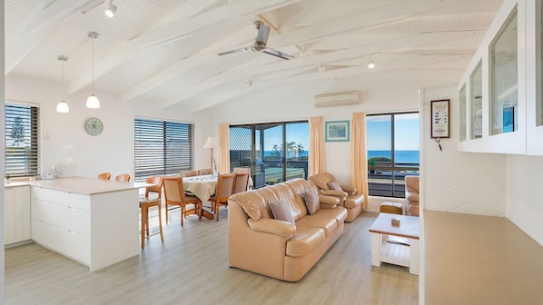 A spacious living area with a large sofa, dining table, and a view of the sea through large windows.