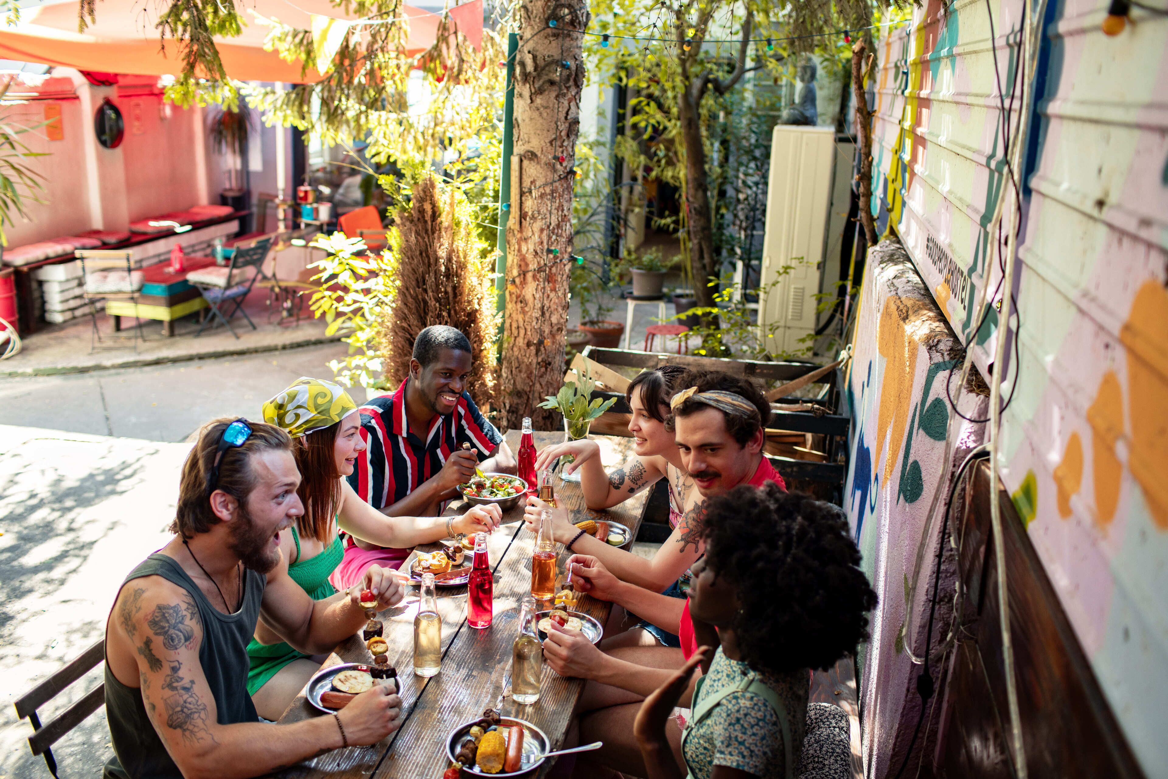 Un groupe de personnes est réuni autour d’une table en bois, à l’extérieur, et déguste divers plats et boissons.