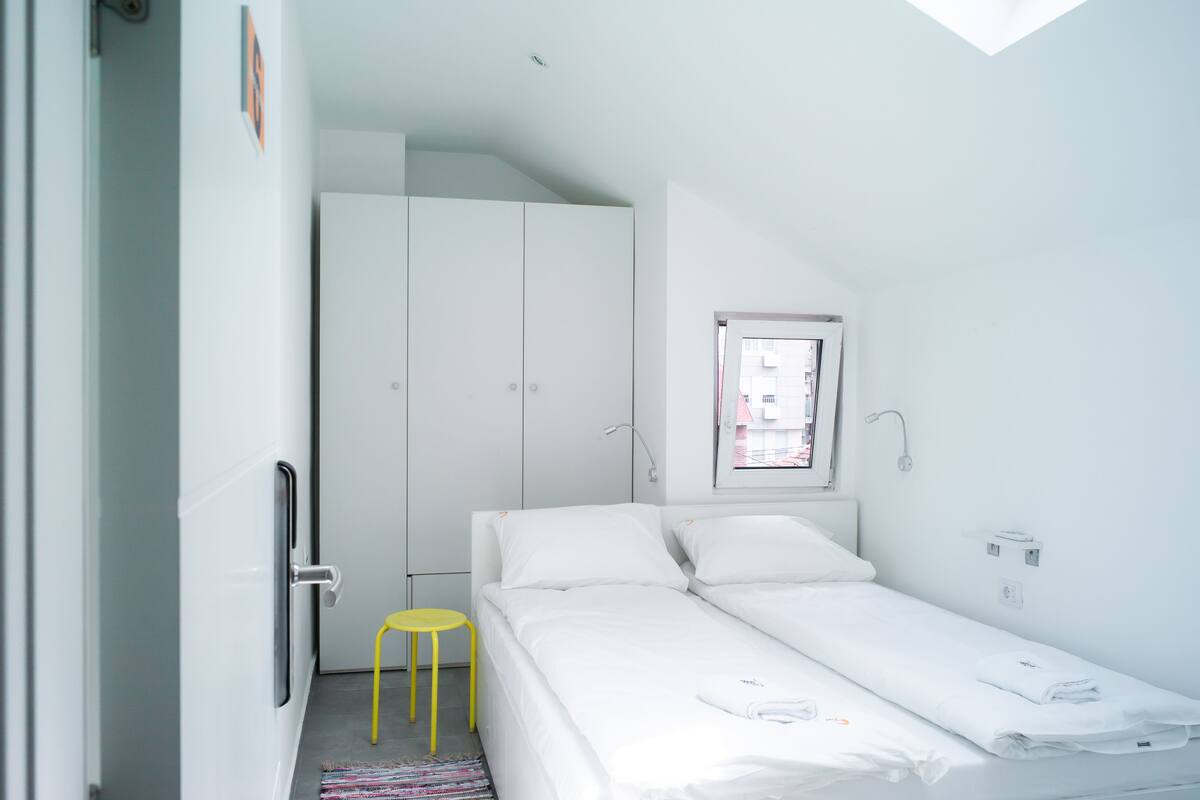 A small, bright room with a bed, a yellow stool, and a wardrobe.