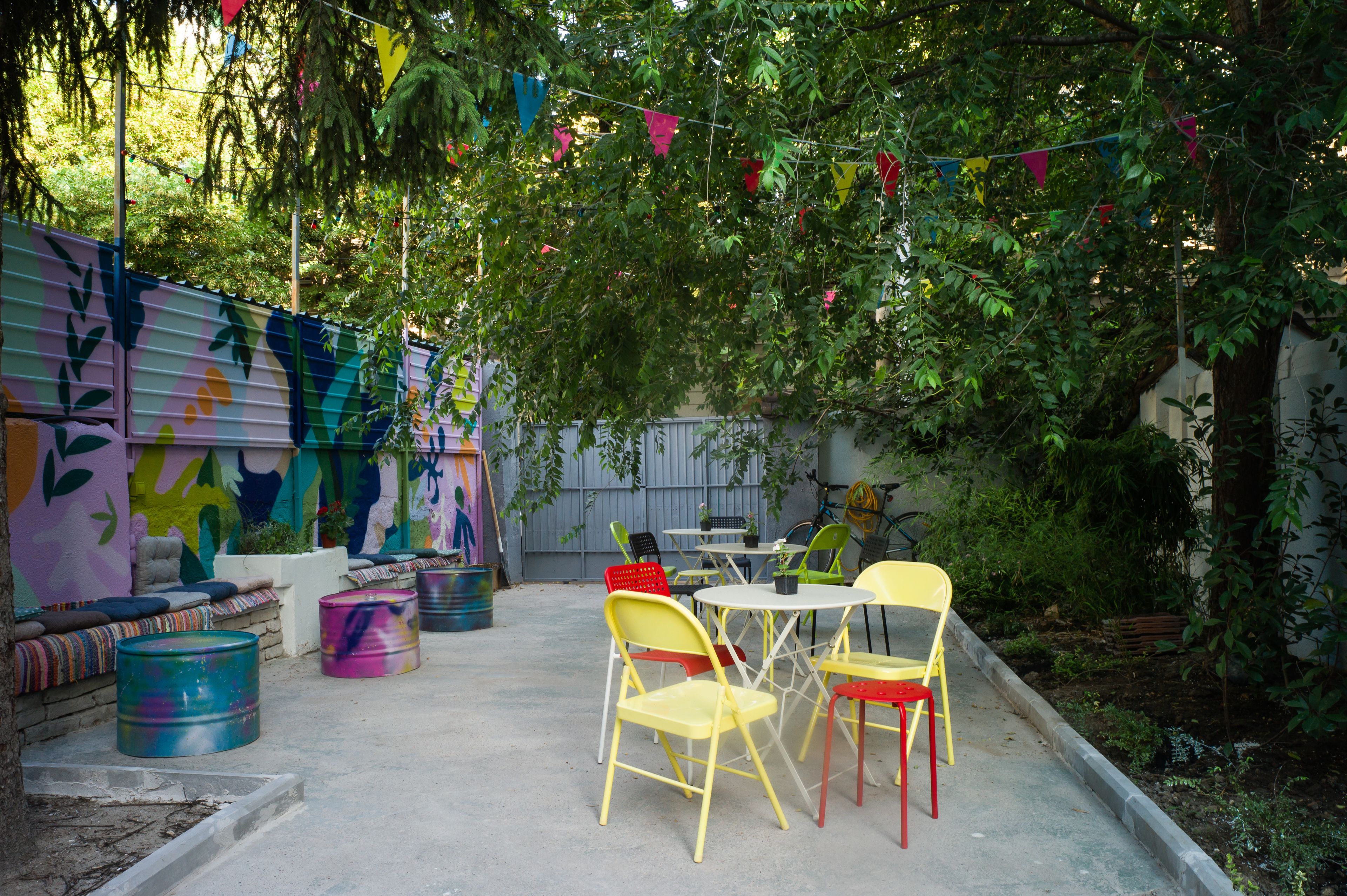 Een terras buiten met kleurrijke stoelen en tafels, omringd door een muurschildering en beplanting.