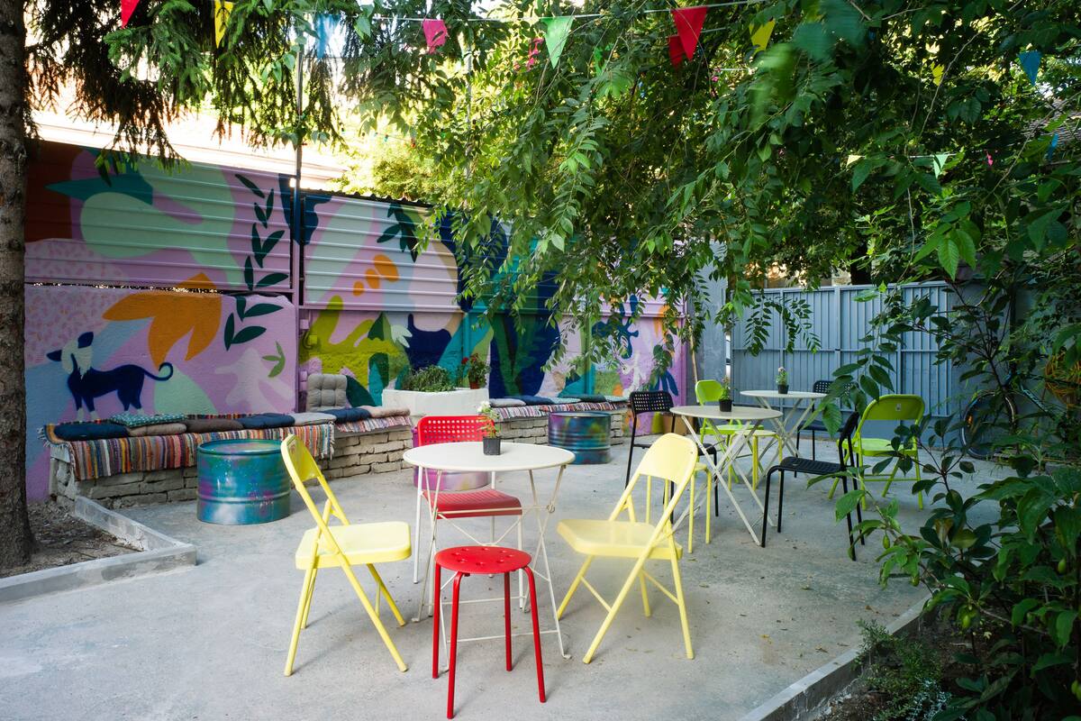 An outdoor patio with colorful chairs and tables, a mural on the wall, and a tree providing shade.