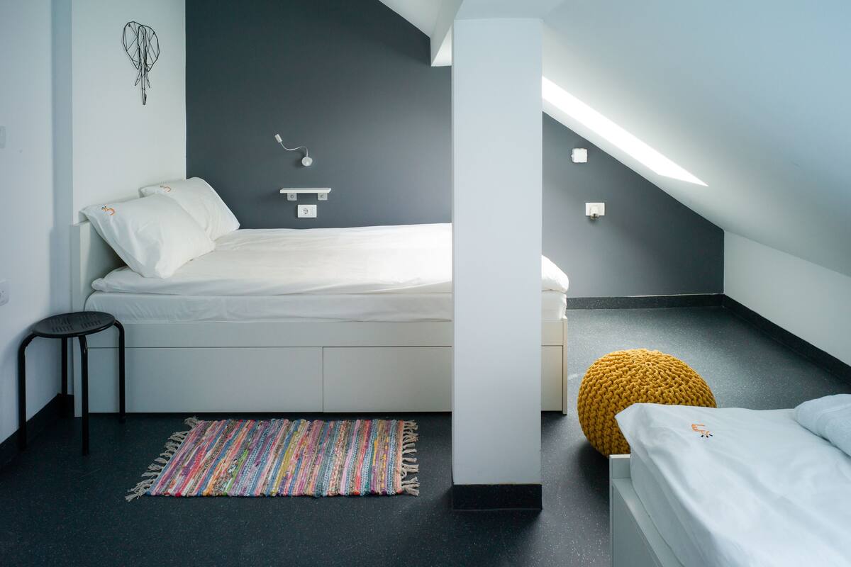 A modern bedroom with a bed, a small stool, a colorful rug, and a unique wall art piece.