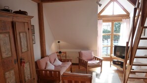 Een gezellige kamer met een houten vloer, een roze bank, een houten stoel, een kast met bloemendesign, een televisie en een raam met gordijnen.
